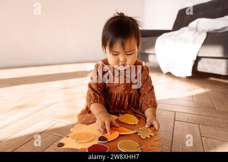 Ein süßes kleines asiatisches Mädchen spielt auf dem Wohnzimmerboden. Ein lustiges Kleinkind in einem braunen Kleid befestigt Blätter an einem Igel. Übungen für die Entwicklung Stockfoto