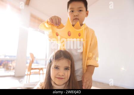 Niedliche Kinder verschiedener Rassen spielen zusammen. Ein kleiner asiatischer Junge hält eine Papierkrone über dem Kopf eines kaukasischen Mädchens. Die Kinder haben Spaß in der Li Stockfoto