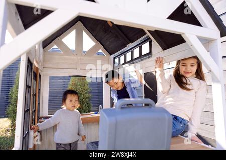 Drei kleine Kinder verschiedener Rassen spielen in einem hölzernen Kinderhaus. Asiatische und kaukasische niedliche Kinder haben gemeinsam Spaß im Garten Stockfoto
