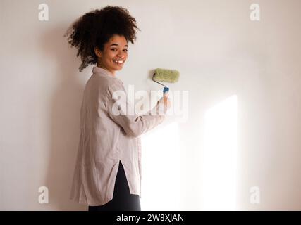 Eine charmante junge Afrikanerin lächelt und hält eine Rolle zum Malen. Attraktives, süßes, gemischtes Mädchen malt die Wände. Renovierung in einem Apartment oder Stockfoto