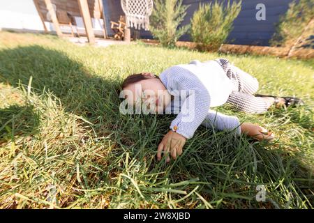 Das süße asiatische Mädchen liegt auf einem grünen Rasen. Ein süßes koreanisches Baby schläft auf dem Gras im Hinterhof des Hauses. Ein kleines Kind in legeren Klamotten spielt Stockfoto