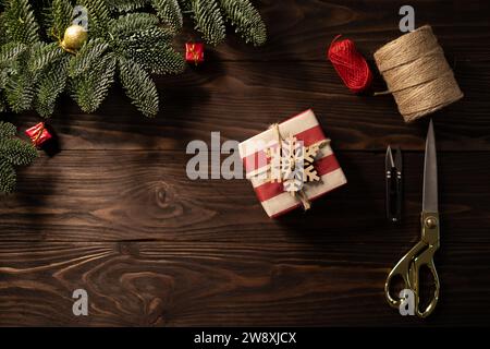 Blick von oben auf das Weihnachtsgeschenk, das in gestreiftes Bastelpapier gewickelt und mit hölzernen Schneeflocken auf hölzernem Hintergrund von edlen Tannenzweigen verziert ist Stockfoto