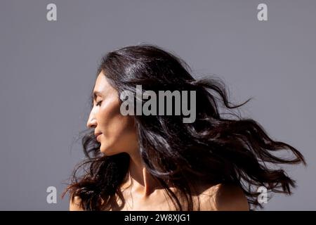 Ein Studio-Modeschuss einer jungen attraktiven Frau mit wunderschönem wellenförmigem Haar und perfekter Haut. Eine charmante Brünette dreht ihren Kopf gegen einen grauen IS Stockfoto