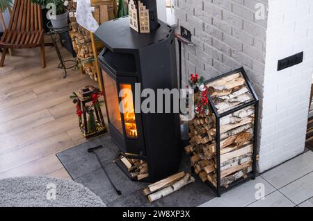 Schwarzer Metallofen, brennender Kamin im weißen festlichen Inneren des Hauses ist für Weihnachten und Neujahr dekoriert, Weihnachtsbaum. Brennholz in t Stockfoto