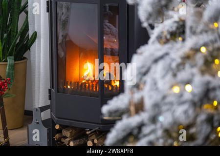 Schwarzer Metallofen, brennender Kamin im weißen festlichen Inneren des Hauses ist für Weihnachten und Neujahr dekoriert, Weihnachtsbaum. Brennholz in t Stockfoto