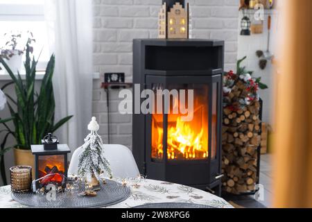 Schwarzer Metallofen, brennender Kamin im weißen festlichen Inneren des Hauses ist für Weihnachten und Neujahr dekoriert, Weihnachtsbaum. Brennholz in t Stockfoto