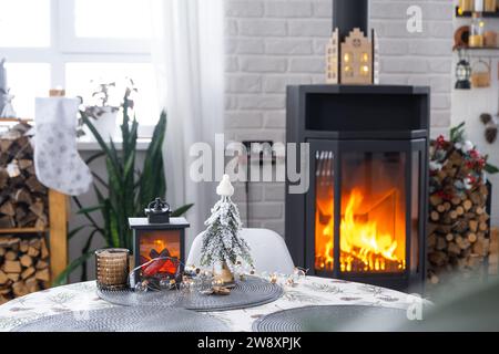 Schwarzer Metallofen, brennender Kamin im weißen festlichen Inneren des Hauses ist für Weihnachten und Neujahr dekoriert, Weihnachtsbaum. Brennholz in t Stockfoto