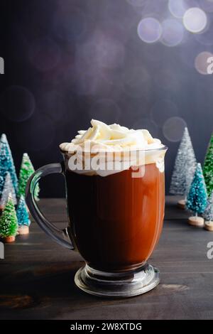 Gewürzter Weihnachtskaffee mit Schlagsahne: Großer Glasbecher mit frisch gebrühtem Gewürzkaffee umgeben von Miniatur-Weihnachtsbäumen Stockfoto