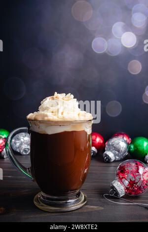 Gewürzter Weihnachtskaffee mit Schlagsahne: Großer Glasbecher mit frisch gebrühtem Gewürzkaffee, umgeben von Weihnachtsschmuck Stockfoto