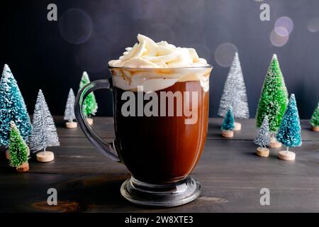 Gewürzter Weihnachtskaffee mit Schlagsahne: Großer Glasbecher mit frisch gebrühtem Gewürzkaffee umgeben von Miniatur-Weihnachtsbäumen Stockfoto