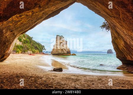 Blick von einer Höhle auf einen Strand und isolierte Felsen im Meer, unter einem bewölkten Himmel, Cathedral Grove, Coromandel, Neuseeland Stockfoto