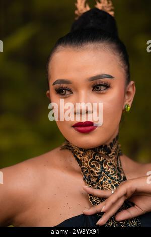 Schöne Augen einer asiatischen Frau in Make-up und rotem Lippenstift, während sie Goldschmuck und schwarzes Haar trägt, während sie vor dem Dschungel steht Stockfoto