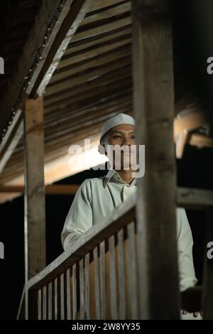 Ein islamischer Mann betet auf dem Balkon, während er in der dunklen Nacht ein weißes muslimisches Kleid im Dorf trägt Stockfoto