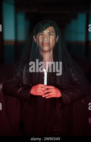 Ein männlicher Teufelsanbeter mit einem transparenten Schleier führt ein gruseliges Ritual durch, indem er nachts eine Kerze in der Hand hält Stockfoto