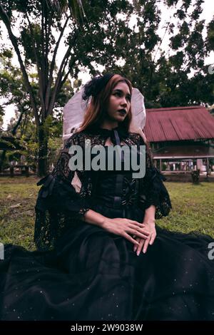 Eine junge Hexe in einem langen schwarzen Kleid und gruseligen Make-up saß nachmittags vor dem Grabstein Stockfoto