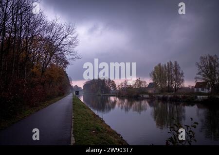 Dieses stimmungsvolle Bild fängt eine Szene am Flussufer bei Sonnenaufgang ein, wo der bewölkte Himmel den Bereich in ein weiches, diffuses Licht hüllt. Ein einsamer Pfad verläuft entlang des Flusses, der den Blick auf eine kleine Figur in der Ferne führt und ein Gefühl von Größe und Einsamkeit verleiht. Die Stille des Wassers reflektiert die Grautöne des Himmels und die Umrisse der Bäume, mit einem Hauch von Sonnenaufgang, der durch die Wolken bricht und einen Hauch von Wärme in einer ansonsten kühlen Palette bietet. Düstere Dämmerung am Riverside Path. Hochwertige Fotos Stockfoto