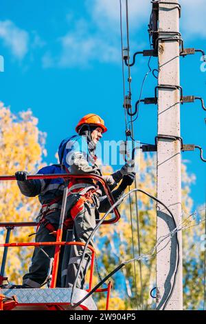 Zwei professionelle Elektriker in Schutzhelmen reparieren Stromleitungen von der Ladestation des Schaufelwagens. Ansicht von unten. Elektriker wechseln Kabel an Straßenbeleuchtungsmasten. Stockfoto