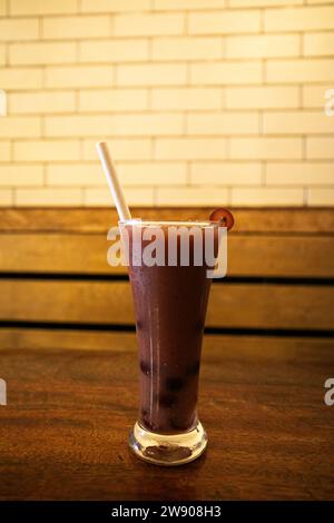 Ein Nahfoto eines Glases lila Flüssigkeit, wahrscheinlich Traubensaft, mit einer einzigen grünen Traube darüber. Das Glas sitzt auf einem dunklen Holztisch, und Stockfoto
