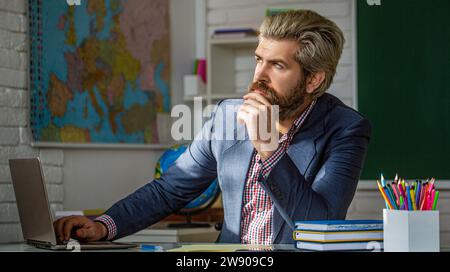 Ein Lehrer mit Laptop in der Klasse sitzt am Tisch, an der Tafel im Klassenzimmer. Lehrer nachdenklich im Klassenzimmer der Grundschule Stockfoto