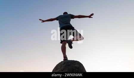 Silhouette eines Mannes, der bei Sonnenuntergang auf einem Felsen im Freien Yoga praktiziert, Konzept des Gleichgewichts und Beruhigung von Geist und Körper Stockfoto