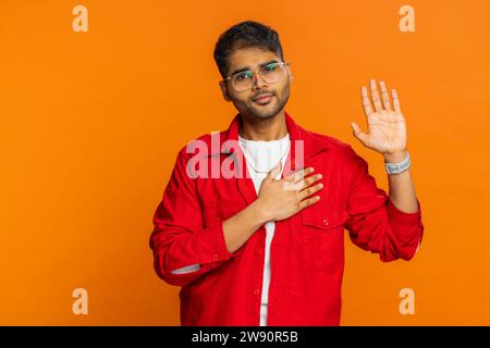 Ich schwöre, ehrlich zu sein. Ein ehrlicher, verantwortungsvoller Indianer erhebt Hand, um Eid zu leisten, verspricht, ehrlich zu sein, die Wahrheit zu sagen, Hand auf Brust zu halten, Gerechtigkeit. Arabianer isoliert auf orangenem Studiohintergrund Stockfoto