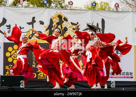 Ein Team japanischer yosakoi-Tänzer tanzt auf der Bühne in roten Röcken und langärmligen Yukata-Tuniken, während sie Naruko und Klatscher halten. Kyusyu Gassai Festival. Stockfoto