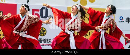 Ein Team japanischer yosakoi-Tänzer tanzt auf der Bühne in roten Röcken und langärmligen Yukata-Tuniken, während sie Naruko und Klatscher halten. Kyusyu Gassai Festival. Stockfoto