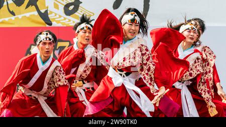 Ein Team japanischer yosakoi-Tänzer tanzt auf der Bühne in roten Röcken und langärmligen Yukata-Tuniken, während sie Naruko und Klatscher halten. Kyusyu Gassai Festival. Stockfoto