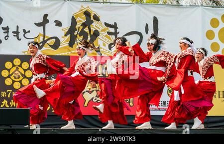 Ein Team japanischer yosakoi-Tänzer tanzt auf der Bühne in roten Röcken und langärmligen Yukata-Tuniken, während sie Naruko und Klatscher halten. Kyusyu Gassai Festival. Stockfoto