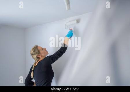 Blonde alleinerziehende Frau, die die Wand ihrer Wohnung mit einer großen Malerwalze in ihrer neuen Wohnung malt Stockfoto