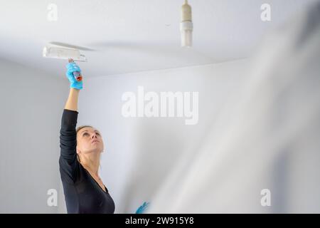 Blonde alleinstehende Frau, die die Decke ihrer Wohnung mit einer großen Malerwalze in ihrer neuen Wohnung malt Stockfoto