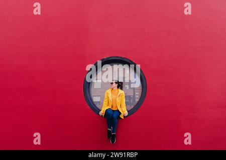 Frau in gelbem Regenmantel sitzt in Bullauge in roter Wand Stockfoto