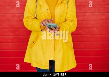 Frau in gelbem Regenmantel mit Smartphone vor roter Wand Stockfoto