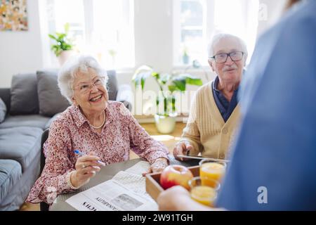 Glückliches Seniorenpaar mit medizinischem Fachpersonal, das ein Tablett mit Apfel und Gläser Saft zu Hause hält Stockfoto