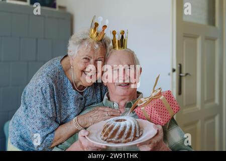 Glückliche Seniorin mit einem Mann, der zu Hause Geburtstagskuchen hält Stockfoto