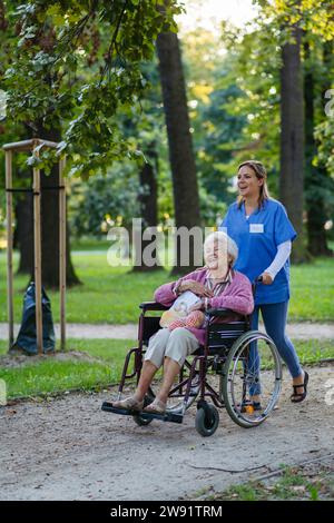 Glückliche Gesundheitsfachkraft, die mit einer älteren Frau im Rollstuhl im Park läuft Stockfoto