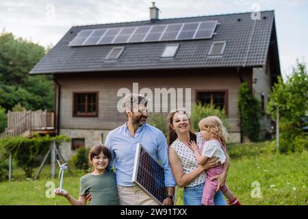 Junge Familie, die vor ihrem Familienhaus mit Solarpaneelen auf dem Dach steht Stockfoto