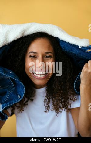 Lächelnde Frau, die eine Jeansjacke auf dem Kopf vor gelbem Hintergrund hält Stockfoto
