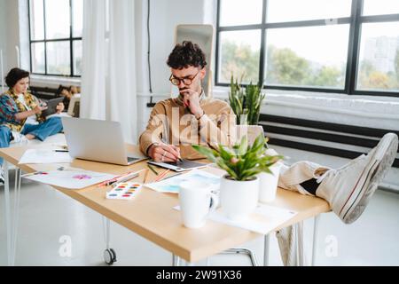 Aufmerksamer Illustrator, der mit den Füßen nach oben am Schreibtisch im Büro arbeitet Stockfoto