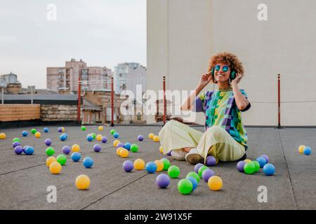Lächelnde junge Frau, die Musik hört, sitzt im Querbein zwischen bunten Bällen auf der Terrasse Stockfoto