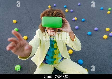 Junge Frau mit VR-Brille, die auf der Terrasse inmitten bunter Bällen sitzt Stockfoto