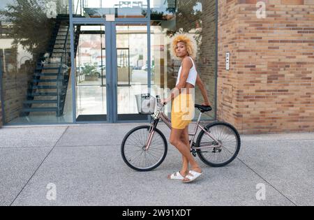 Junge Transgender-Frau, die mit dem Fahrrad auf dem Fußweg vor dem Gebäude steht Stockfoto