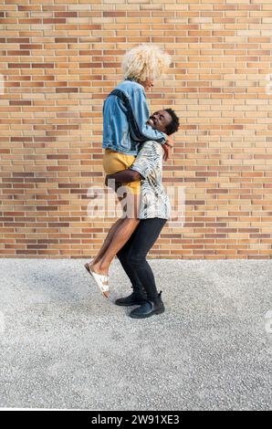 Ein Freund hebt Transgender-Frau vor einer Ziegelmauer auf Stockfoto