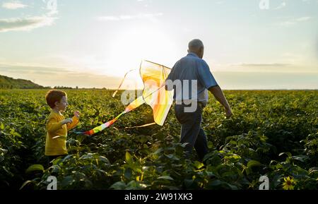 Großvater hält Drachen und geht mit Enkel auf dem landwirtschaftlichen Feld Stockfoto