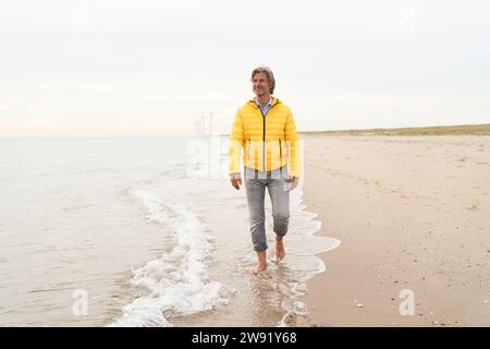 Senior-Mann mit gelber Jacke und am Strand spazieren Stockfoto