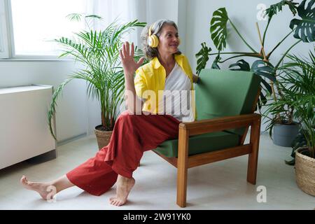 Lächelnde Frau mit kabellosen Kopfhörern, die zu Hause auf dem Stuhl sitzt Stockfoto