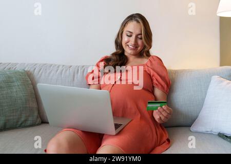 Schwangere Frau beim Online-Shopping über Kreditkarte und Laptop, die zu Hause auf dem Sofa sitzt Stockfoto