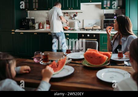 Mutter und Töchter essen am Tisch mit Mann, der Wassermelone in der Küche schneidet Stockfoto