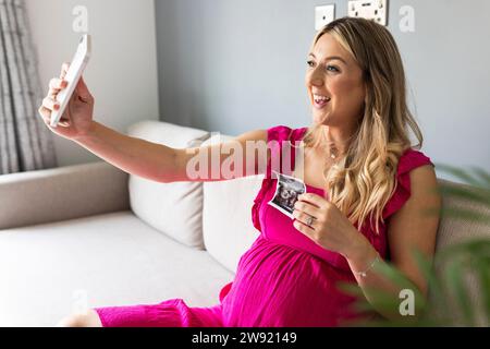 Lächelnde schwangere Frau mit Ultraschallfoto und Selfie durch Smartphone zu Hause Stockfoto