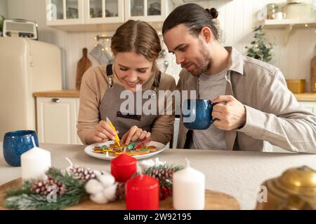 Lächelnde Frau, die Kekse mit einem Mann dekoriert, der in der Küche einen Becher hält Stockfoto